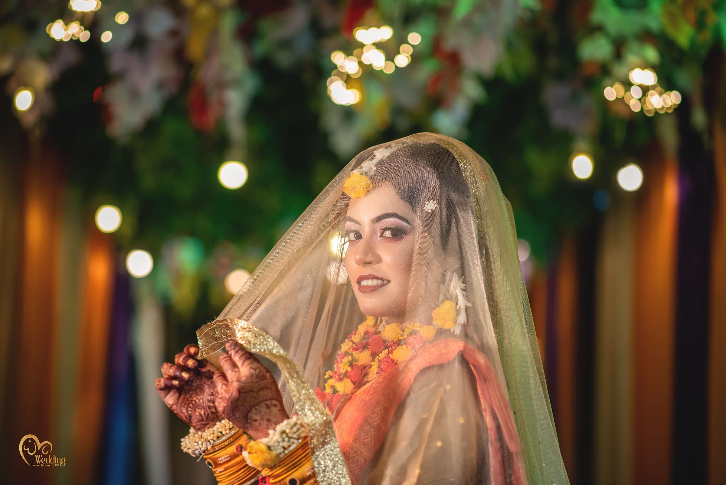 Wide frame of a bride in a sheer veil surrounded by glowing lights and flowers